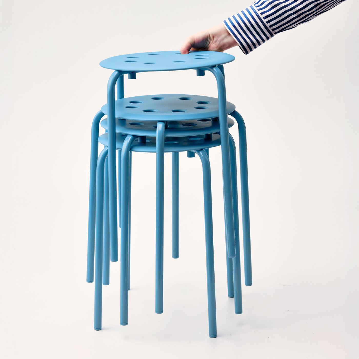 A person is lifting a stack of three vibrant blue MARIUS stools with circular seats and slender legs. 