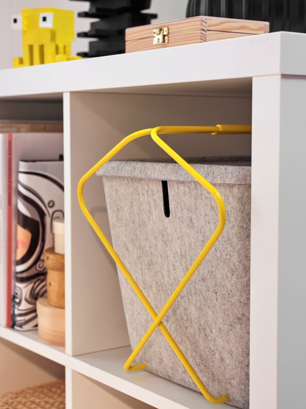 A close-up of a grey and yellow BRÄNNBOLL basket being stored within a white KALLAX shelving unit.