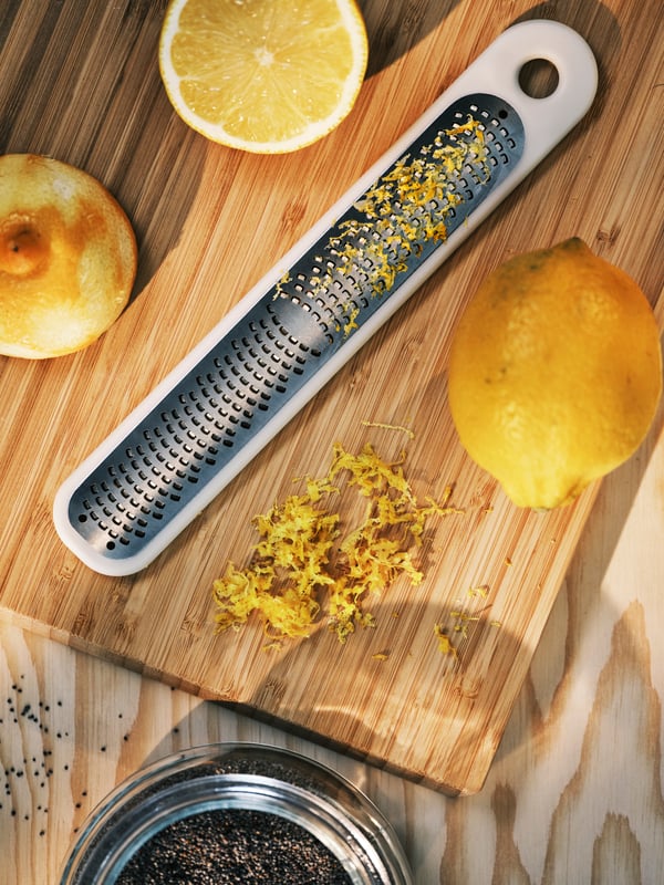 An APTITLIG chopping board in bamboo with an UPPFYLLD grater in off-white, some lemons, and a jar with lid in glass.