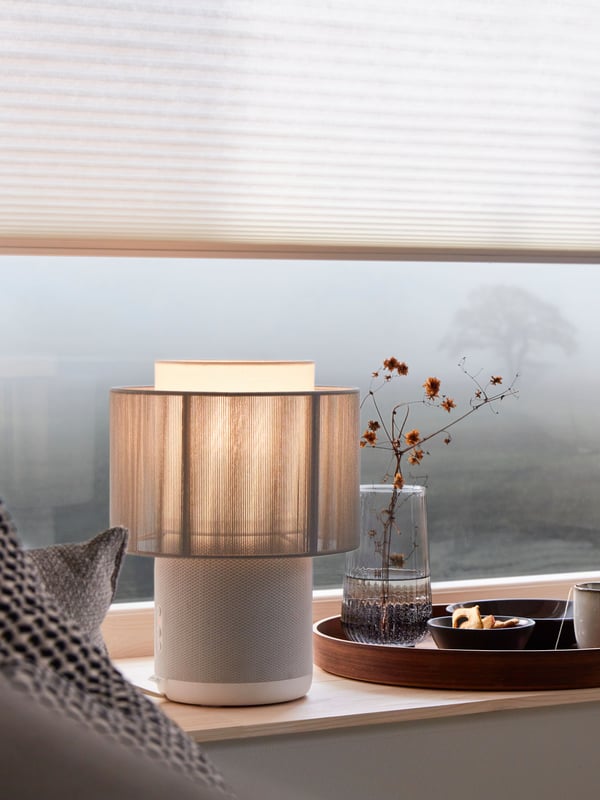 Une lampe enceinte WiFi SYMFONISK éclaire un appui de fenêtre donnant sur un paysage brumeux sous un store alvéolaire PRAKTLYSING.