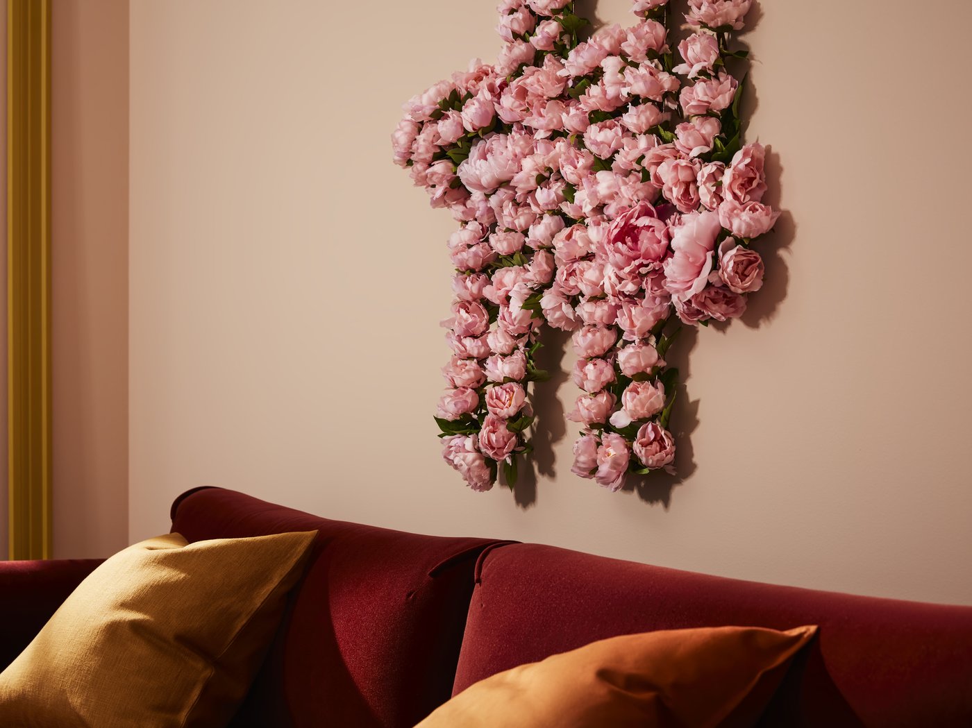 Une décoration murale réalisée avec des fleurs artificielles SMYCKA roses, accrochée au-dessus d’un canapé en velours rouge garni de coussins jaunes.