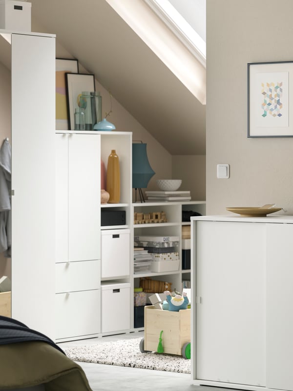 A white VIHALS wardrobe combination under a sloped roof with vases, pictures, storage boxes and other objects on the shelves.