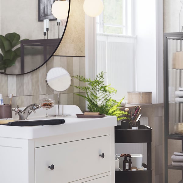 A bright bathroom with a window has a white HEMNES washstand with a round mirror above it and a black trolley next to it.
