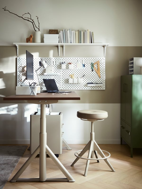 A brown/beige sit/stand desk with a work lamp, laptop and monitor on top and a TROLLBERGET active sit/stand support in front.