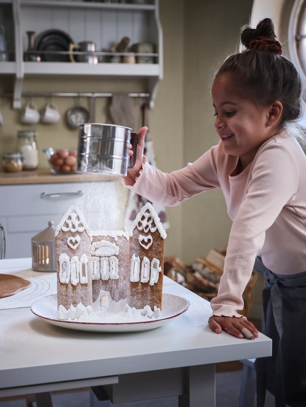 En barn använder en mjölsikt för att dekorera ett pepparkakshus.