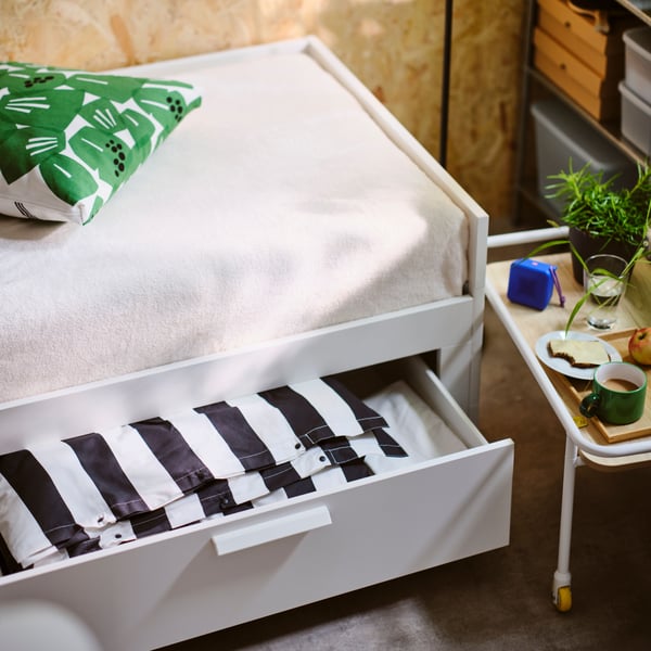 Gros plan sur un lit BRIMNES avec boîte de rangement ouverte dessous, révélant une housse de couette à rayures noir/blanc pliée.