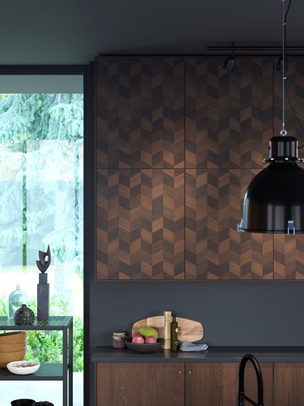 Wooden cabinets on a wall with a chevron pattern on the doors, black pendant lamp, a bowl of fruit on the black worktop.