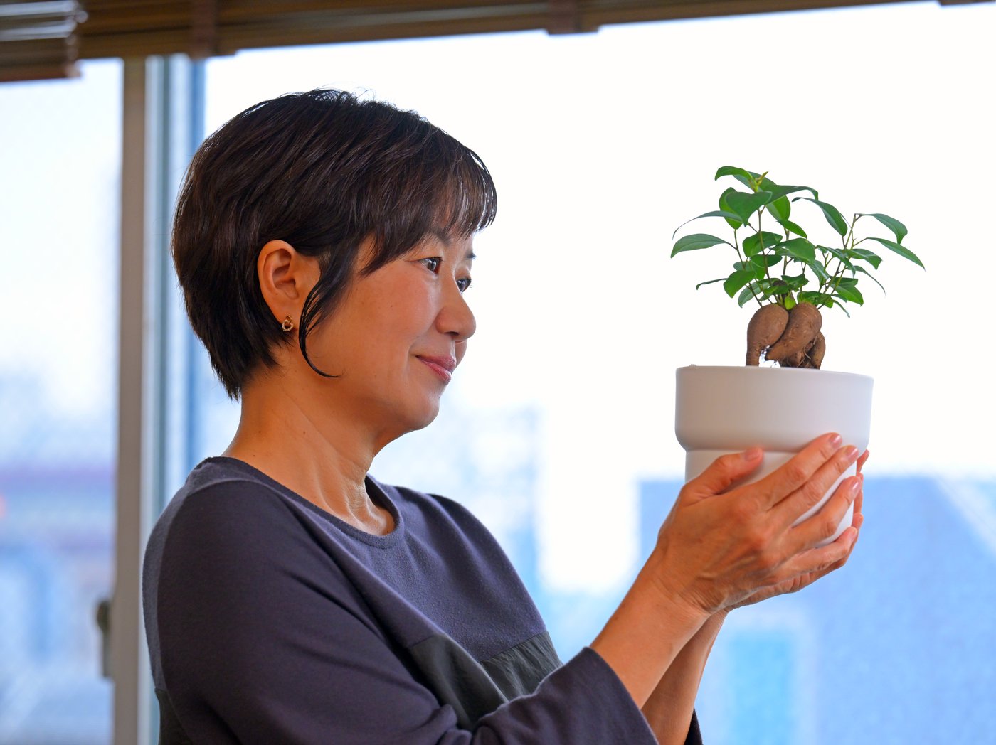 Žena drži belu saksiju u kojoj je malo bonsai drvo.