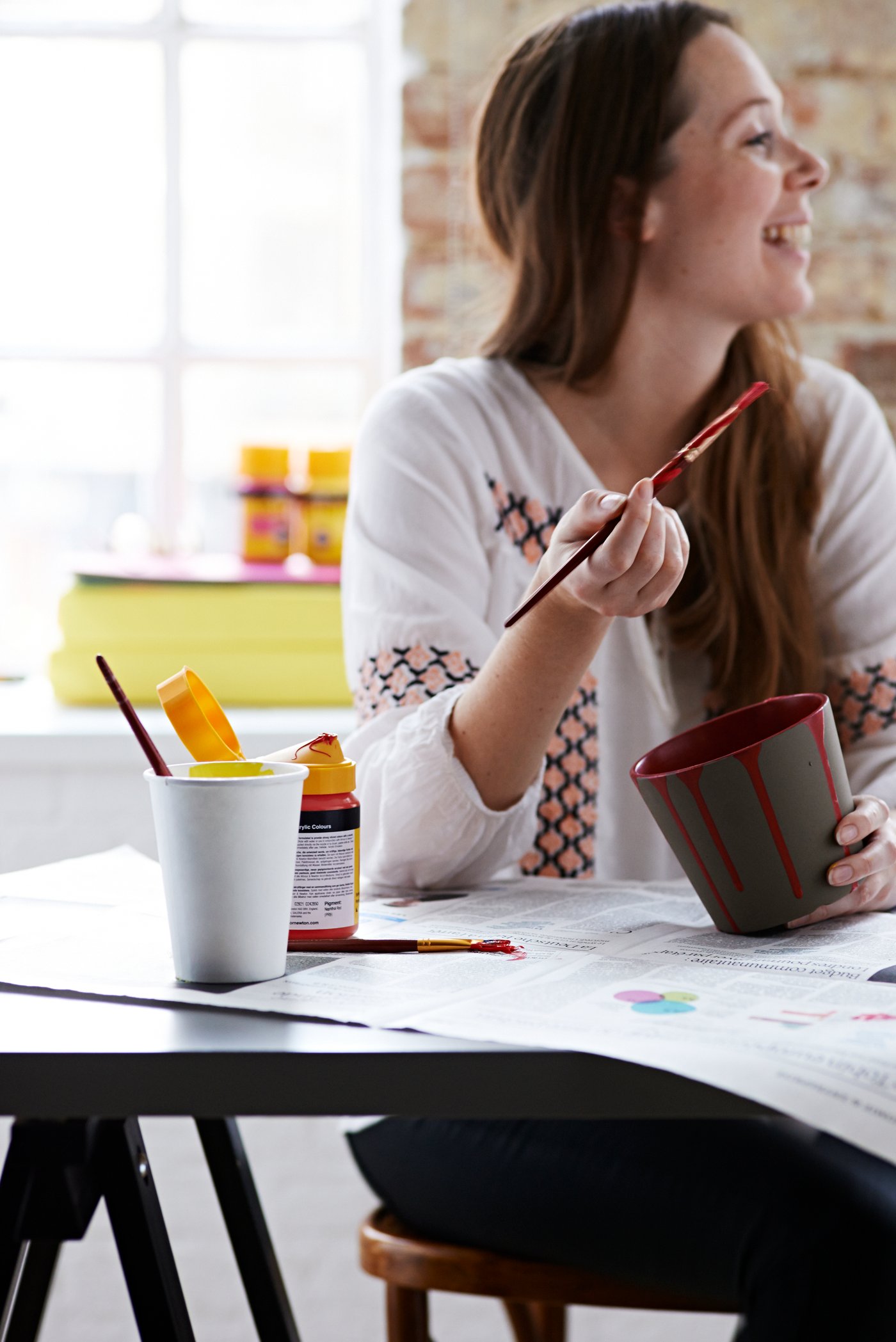 A person sitting at a dining table and painting a grey plant pot with red paint.
