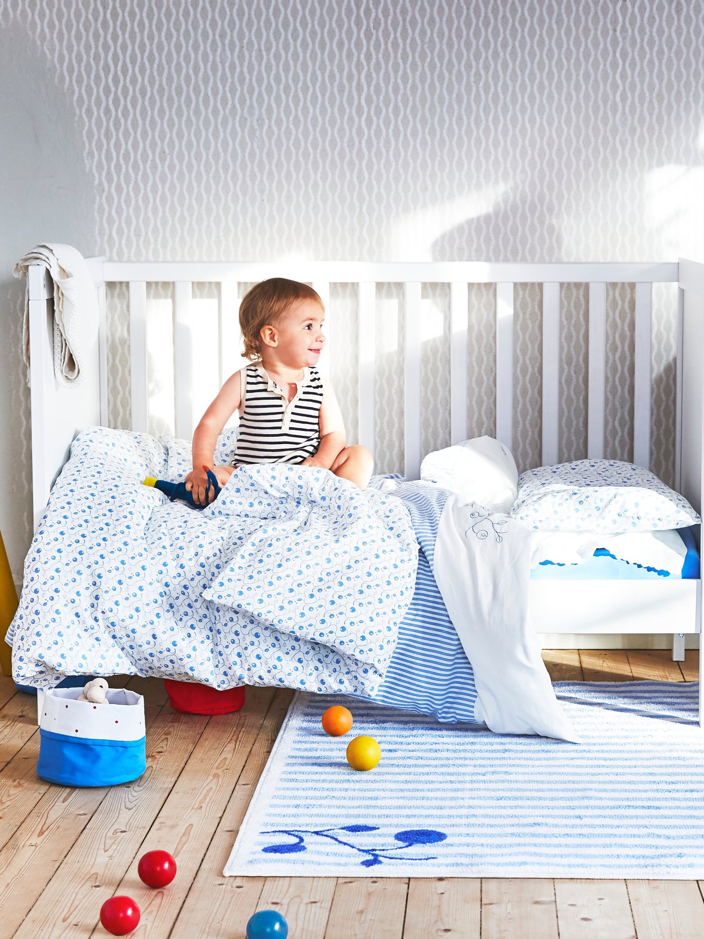 Un petit enfant est assis bien droit sur un lit d'enfant SUNDVIK blanc dont un côté est ouvert, sur lequel sont disposés de nombreux draps et couvertures ornés de motifs de myrtilles.