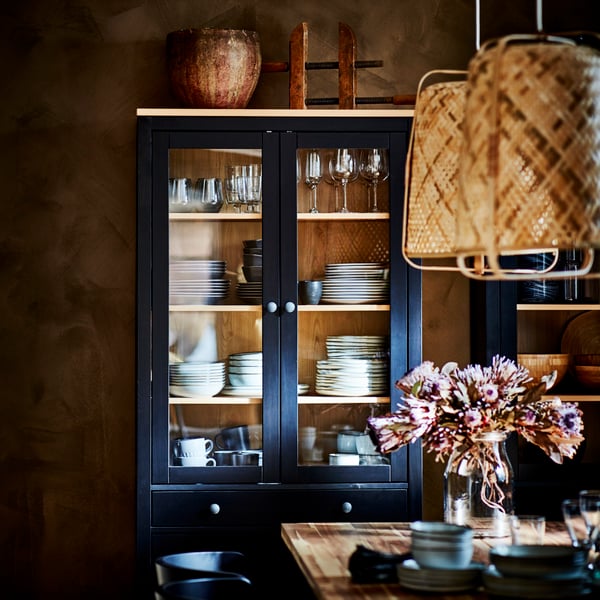A HEMNES glass door cabinet with drawers holding tableware. Two KNIXHULT pendant lamps hang in front over a table.