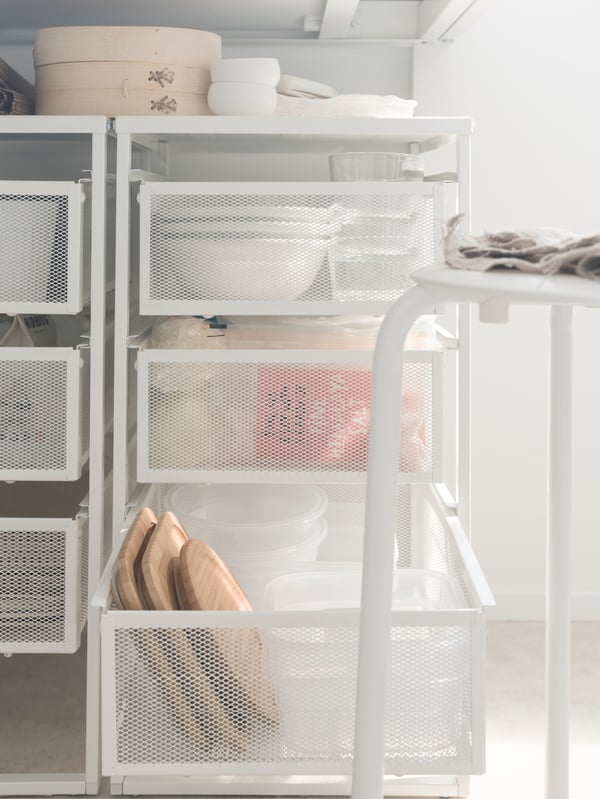 Two white metal mesh LENNART drawer units hold tableware and food storage boxes under a kitchen table, by a white stool.