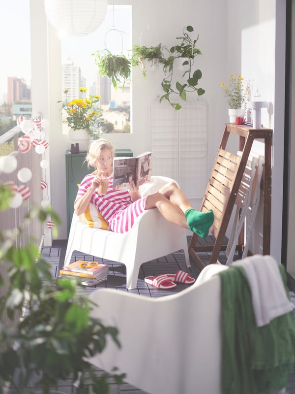A person reading a book in one of two white SKARPÖ outdoor armchairs next to a DJUPÖN folding table on a balcony with plants.