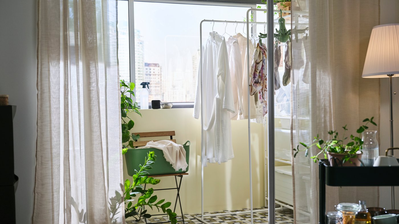 A sunlit balcony with a white clothing rack holding bathrobes and towels, set against a city view. The scene suggests an airy and clean indoor space.