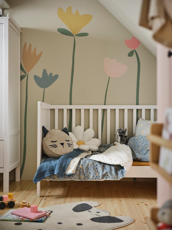 A cot with the side removed covered in blue/white floral pattern GULDVÄVARE bed linen and a BARNDRÖM cushion with pocket.