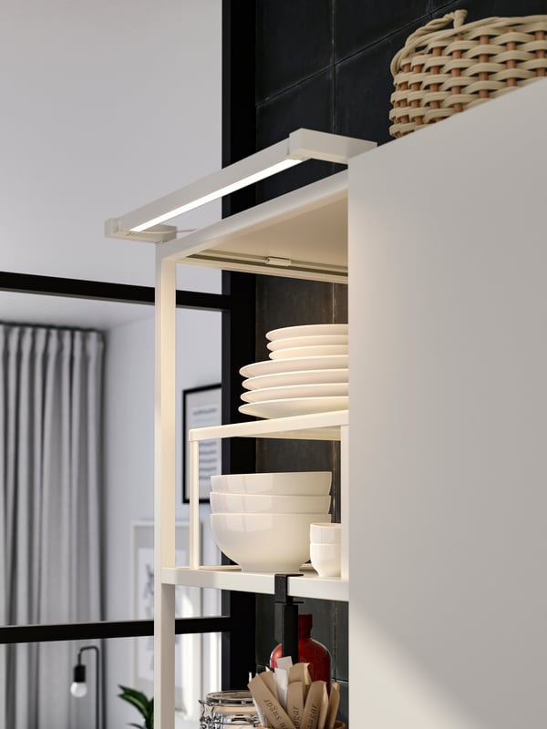 A white SKYDRAG LED worktop lighting strip is attached to a white ENHET wall frame with shelves with white bowls and plates.