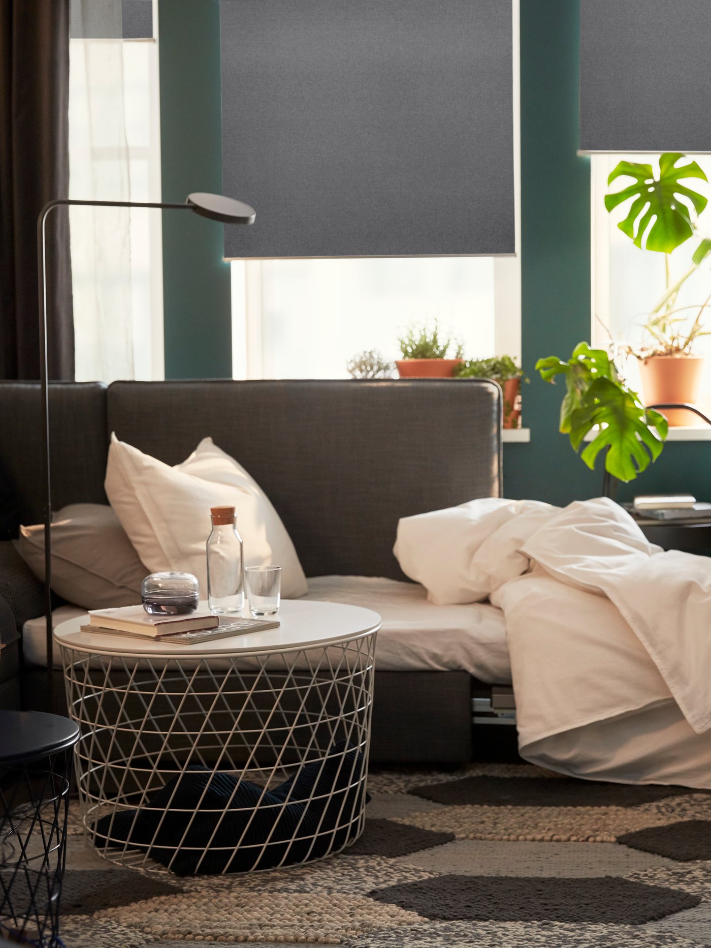 Living room with storage table, floor lamp, sofa, two pillows, potted plants, grey blinds, and rug.
