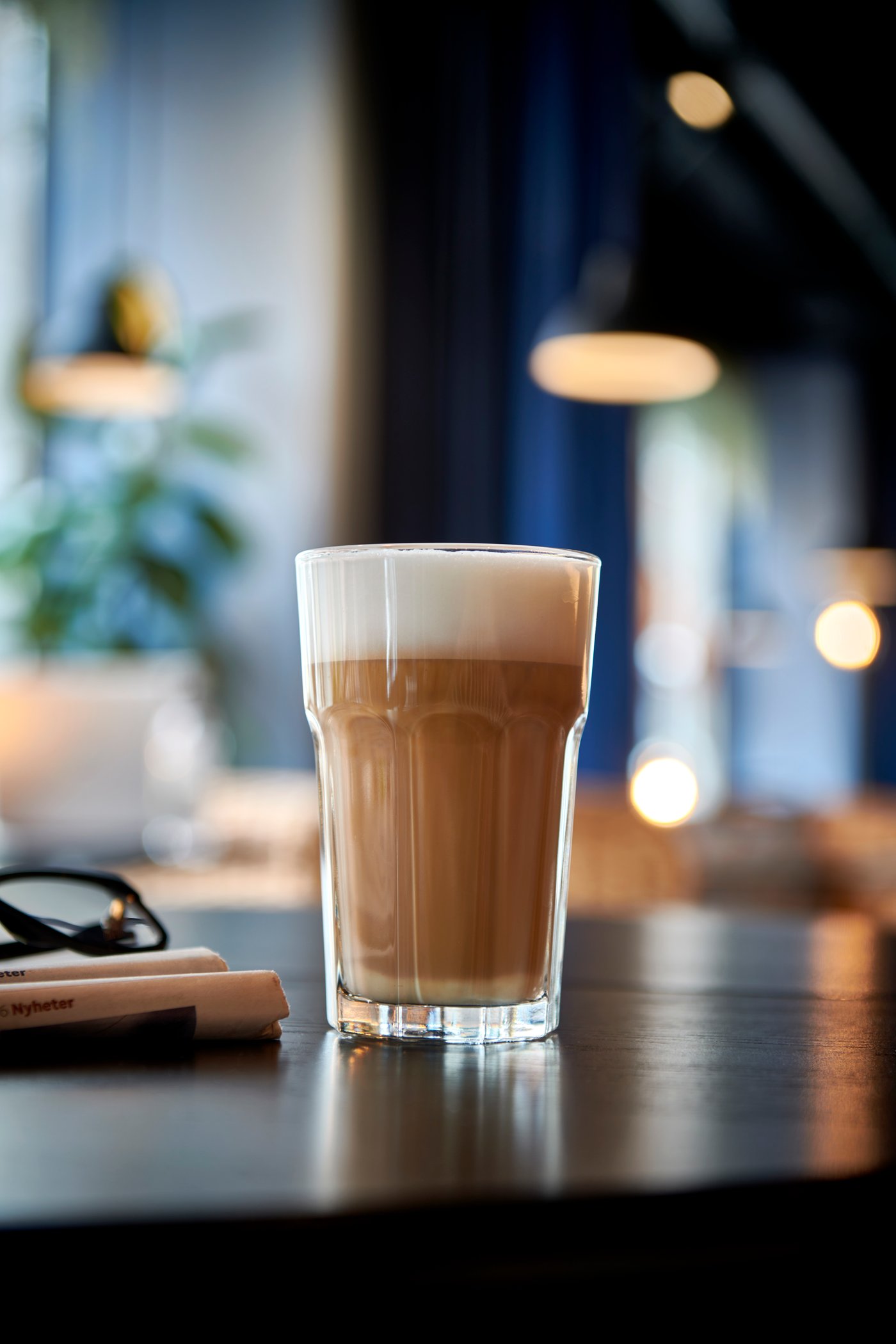 Ett glas latte står på ett mörkt bord i en modern miljö med mjuk belysning. I bakgrunden syns suddiga lampor och en växt, samt en bok och ett par glasögon bredvid glaset.