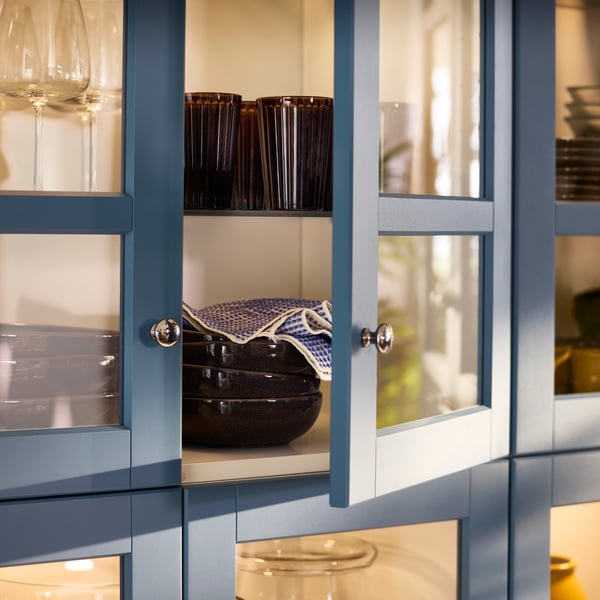 Es ist ein Esszimmerschrank mit LERHYTTAN Vitrinentüren in Blau zu sehen. Auf Böden werden Gegenstände für das Esszimmer aufbewahrt.
