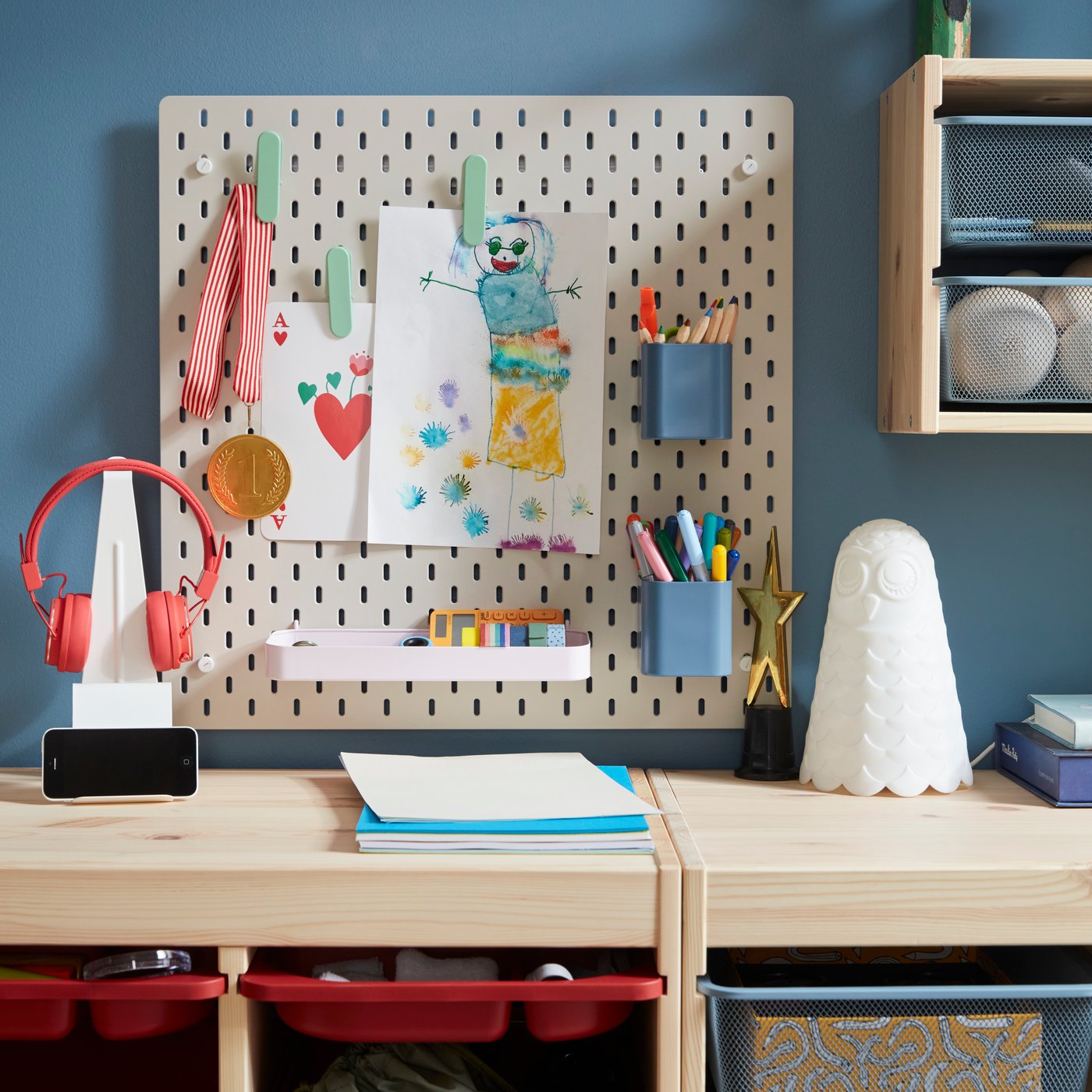 A beige SKÅDIS pegboard with colourful accessories holding pens and drawings placed on a blue wall above storage in pine.
