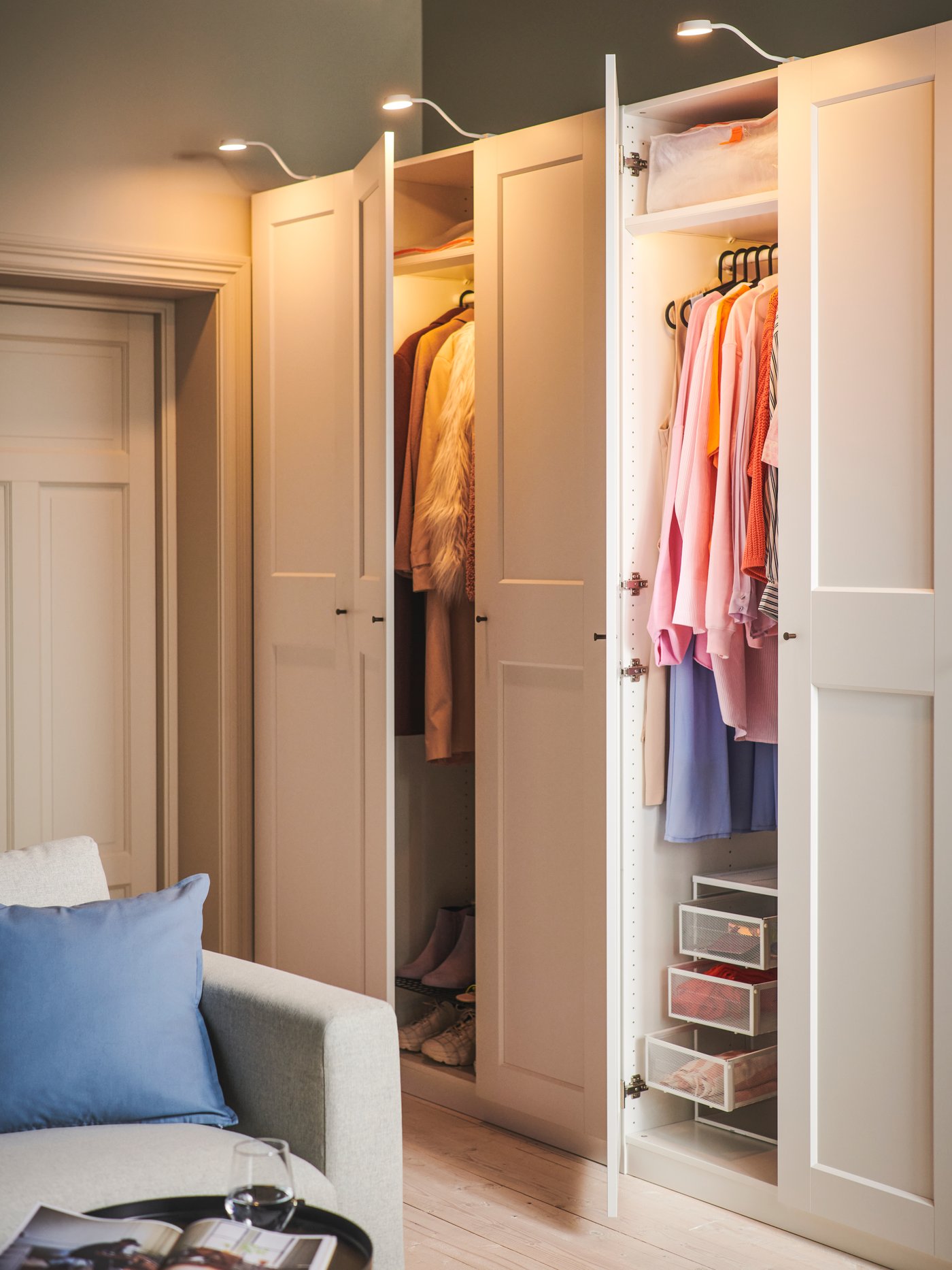 PAX wardrobe in bedroom setting with white hinged doors. There are multiple clothes in the cupboard with stacks of folded clothes. Integrated lighting illuminates the insides of the wardrobe.