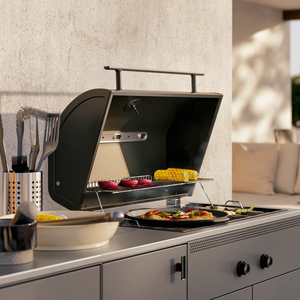 Gros plan sur la zone barbecue d’une cuisine d’extérieur BÅTSKÄR gris foncé. Le couvercle ouvert dévoile une pizza et des légumes.