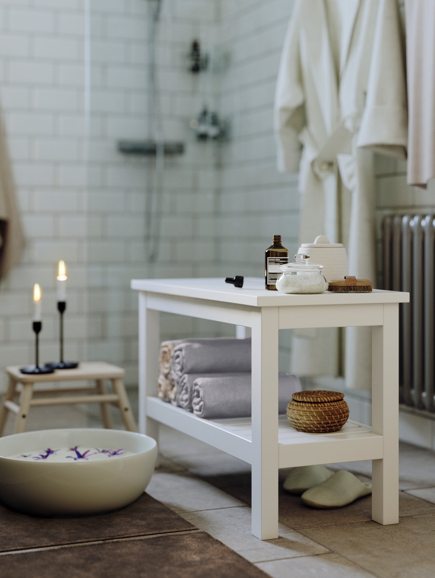 A white HEMNES bench in a white tiled bathroom with rolled-up towels and a box on the lower shelf and containers on top.