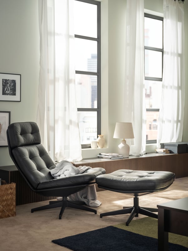 A black-leather HAVBERG swivel armchair and footstool placed on a carpeted floor by tall windows in a softly sunlit room.