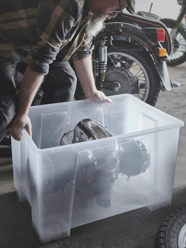 U garaži u kojoj se nalaze motocikli, osoba gura SAMLA kutiju na točkiće i drži motor motocikla.