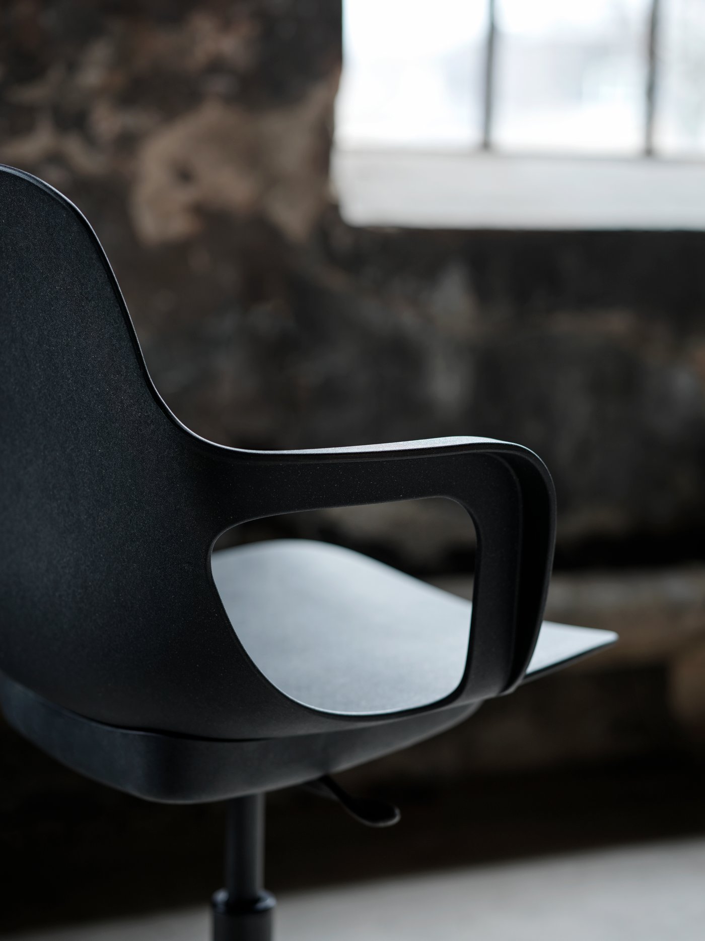 An anthracite ODGER swivel chair standing in a dimly lit room with rough walls and a large, gridded window.
