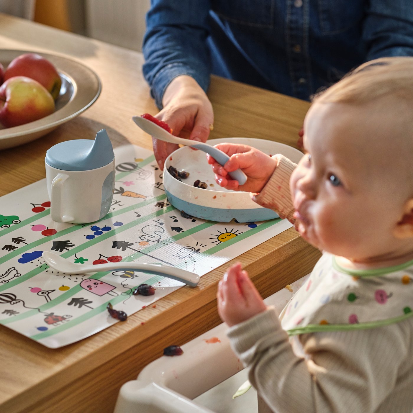 Ett barn håller i en BÖRJA matningssked och sitter i en ANTILOP barnstol vid ett matbord, med en BÖRJA tallrik och mugg på bordet.