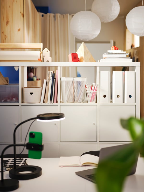 A white KALLAX shelving unit stores multiple things, such as a white RISATORP baskets and three white KUGGIS magazine files.