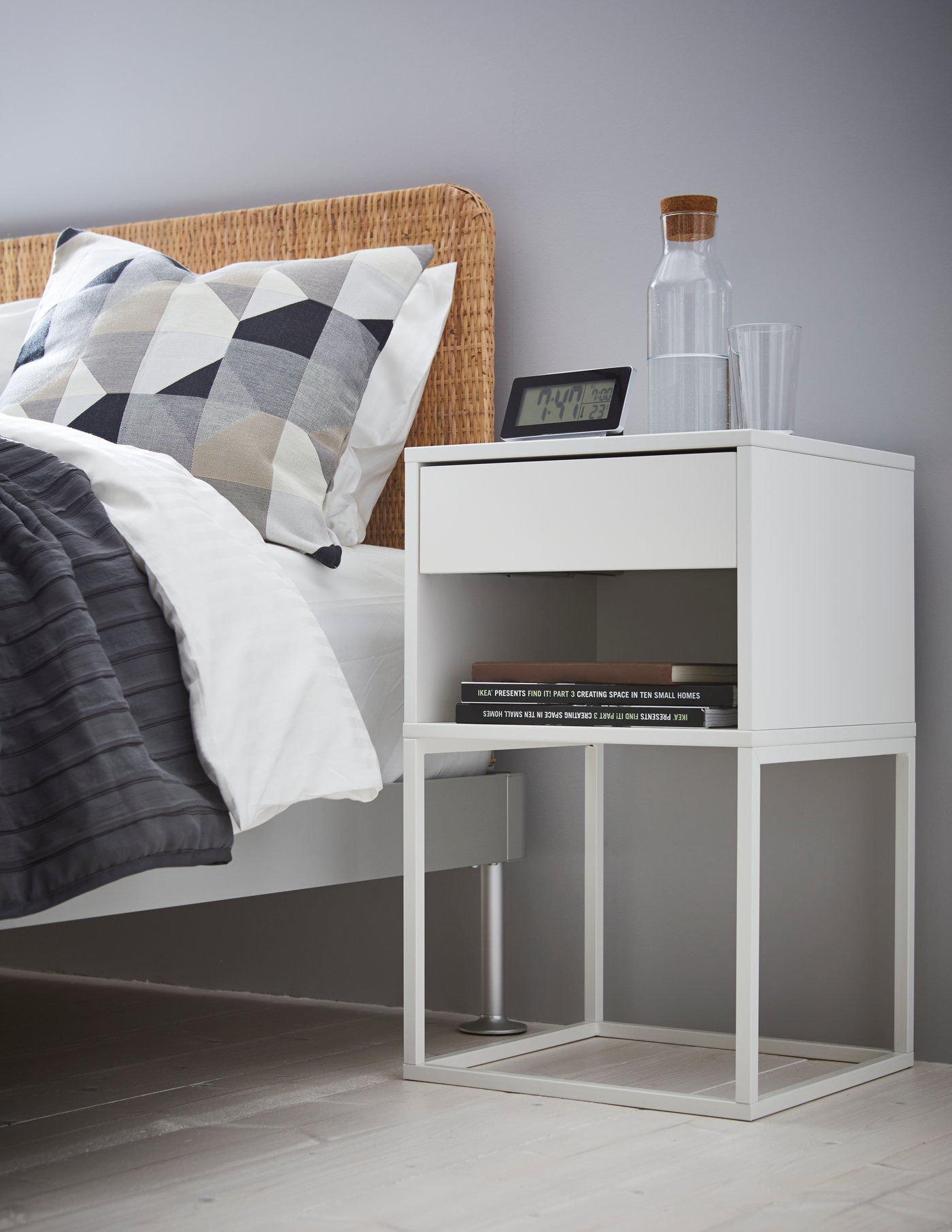 Une table de nuit à côté d’un lit aux draps blancs et accents gris présente une étagère ouverte avec des livres, un tiroir, et sur le dessus une horloge digitale, deux verres et une carafe.