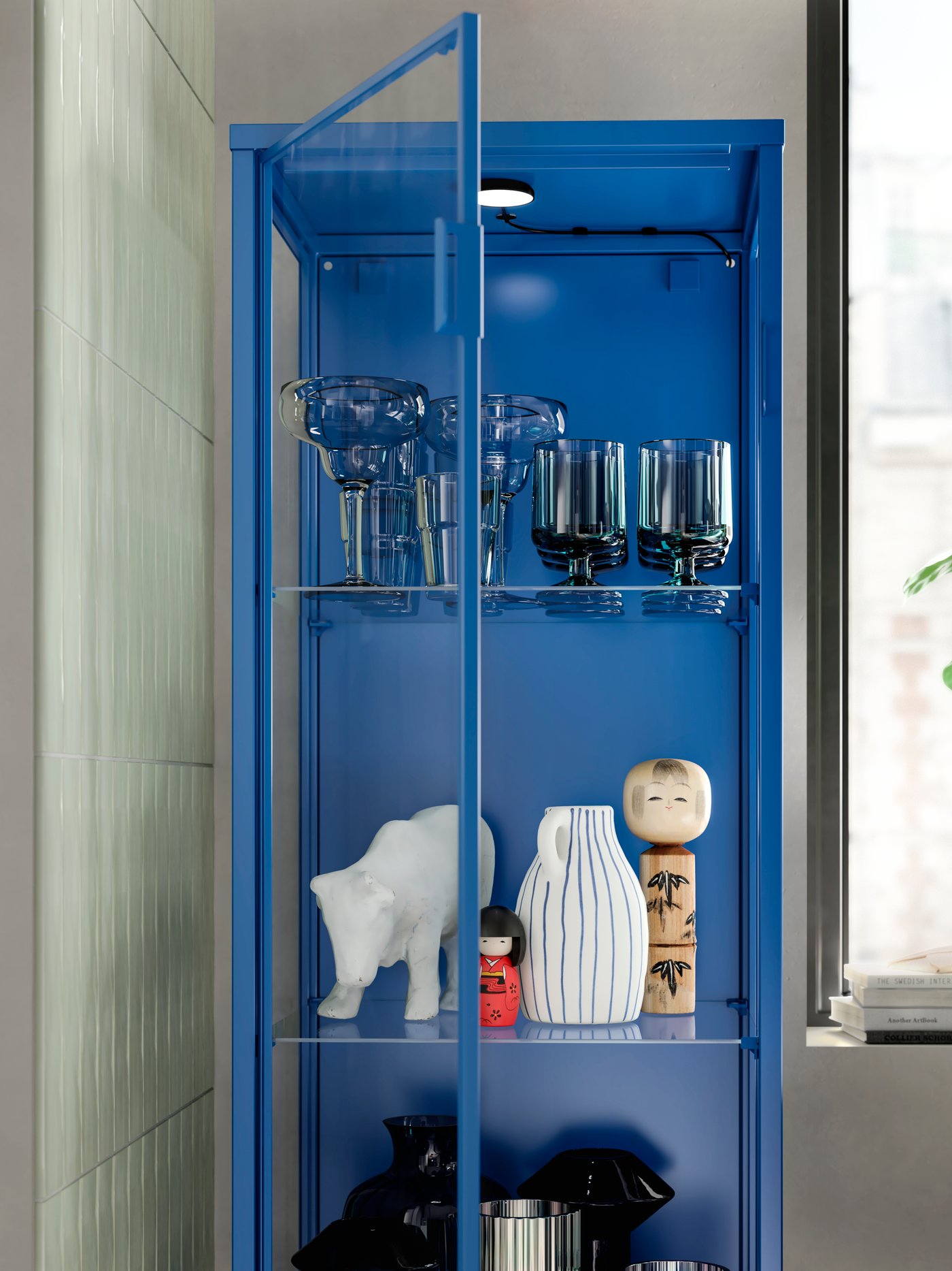 Eine blaue RUDSTA Vitrine steht in einem Wohnzimmer neben einem hellen Fenster.