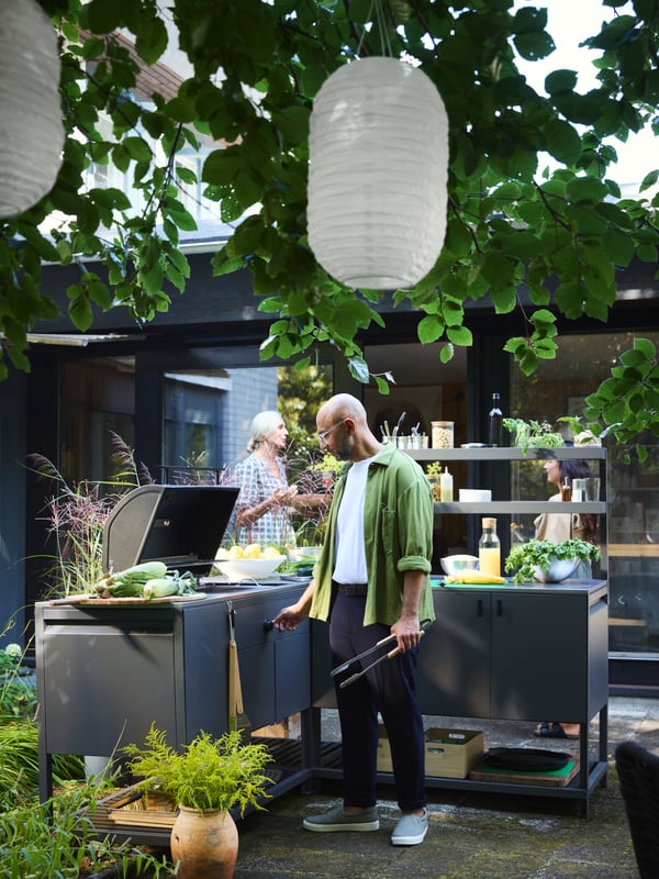 Dans un jardin, un homme utilise le barbecue de la cuisine d’extérieur BÅTSKÄR et deux femmes discutent en arrière-plan.