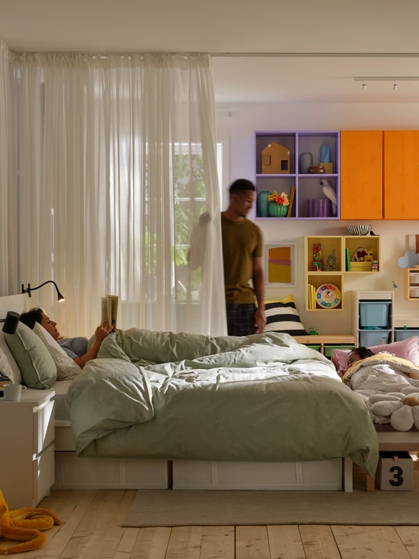 Two adults and a child in a room. The child and one adult lie in separate beds. The other adult is standing by a curtain.
