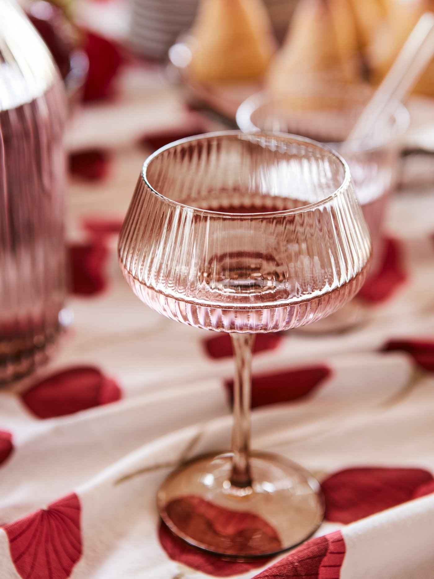 An ANLEDNING champagne coupe in tinted glass with a ridged pattern, holding a drink, placed on a poppy-patterned tablecloth.