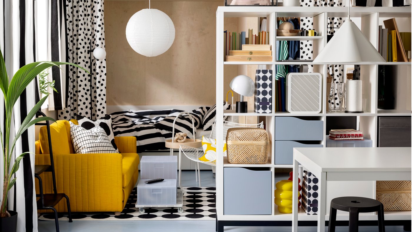 A FRIDHULT sofa-bed stands in a living room area near a white KALLAX shelving unit and a bed with striped bed linen.