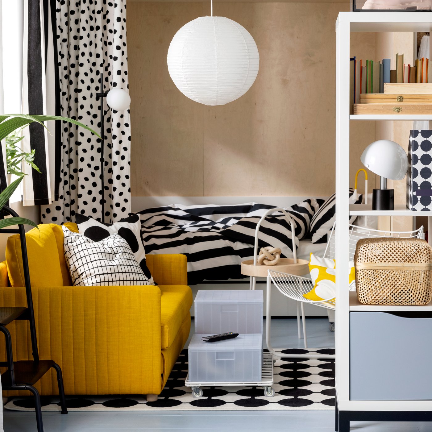 A FRIDHULT sofa-bed stands in a living room area near a white KALLAX shelving unit and a bed with striped bed linen.