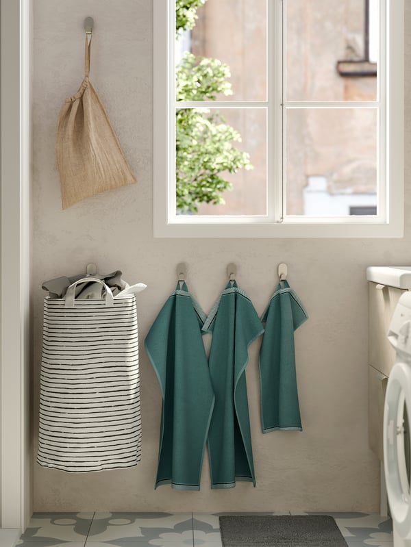 Three light blue towels, a white and black striped laundry basket and a beige linen bag all hung on hooks on the wall.