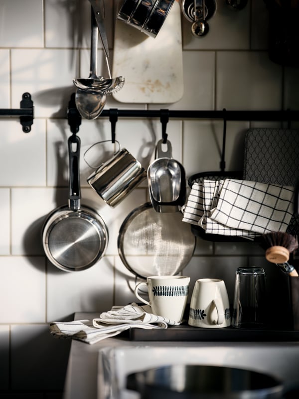 A white-tiled kitchen wall with utensils hanging from a black HULTARP rail, including a RINNIG tea towel and an ANNONS pot.