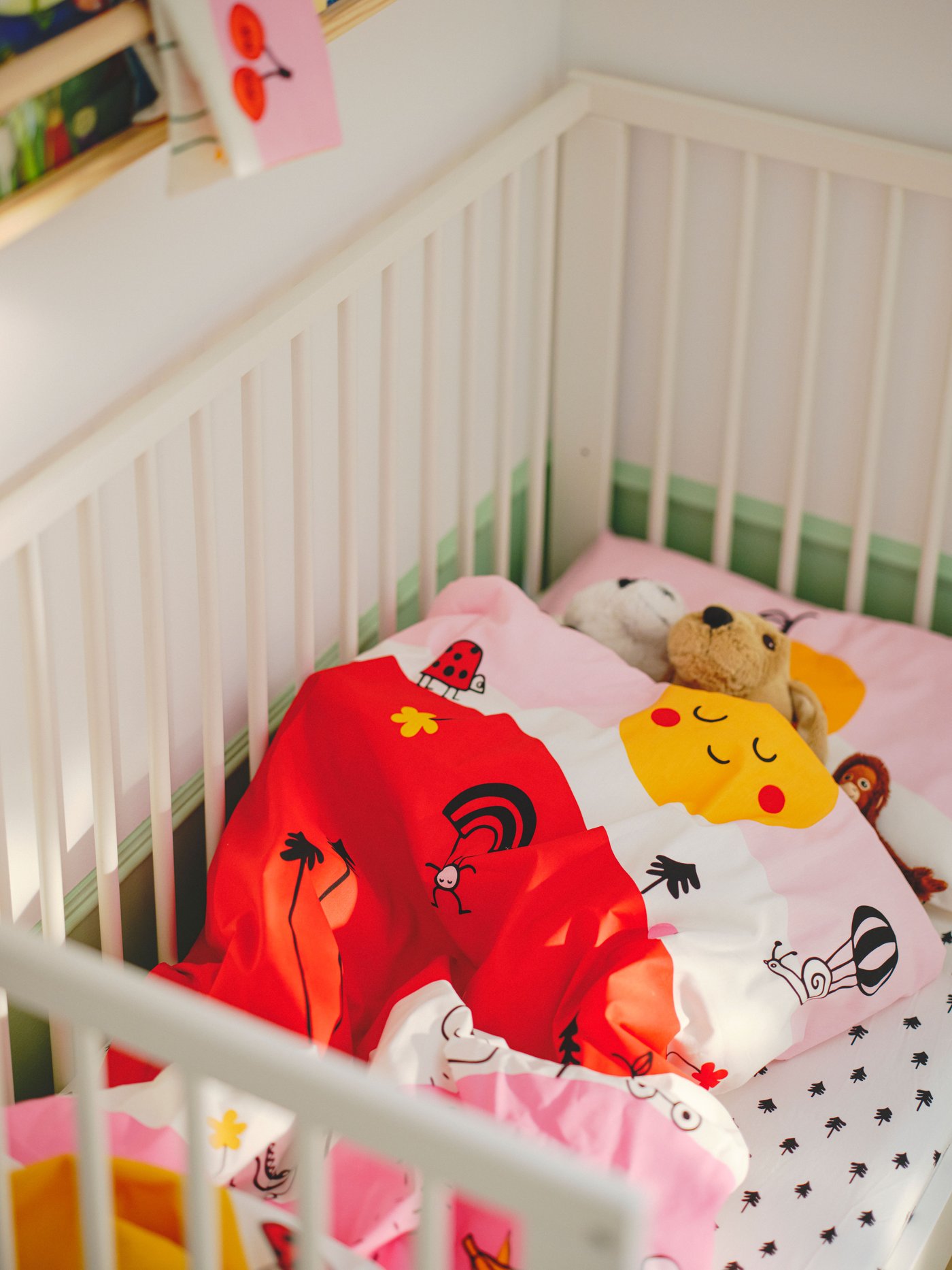 A white GULLIVER cot made up with multicolour/striped GRÖNFINK bed linen and with three soft toys tucked up in the duvet.