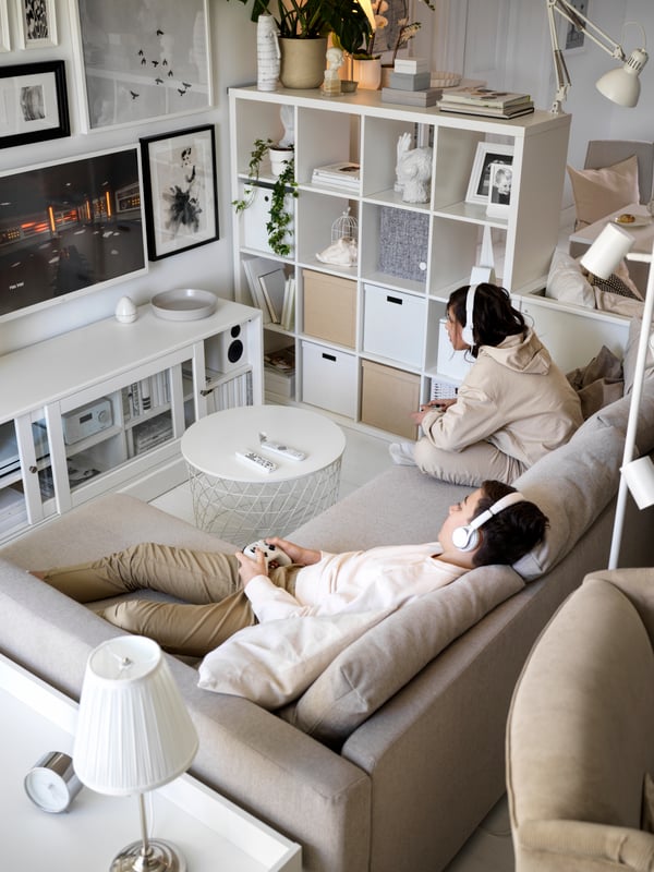 Two young people chilling on a sofa and playing a video game. The seating area is separated from the sleeping area by a KALLAX shelf unit.