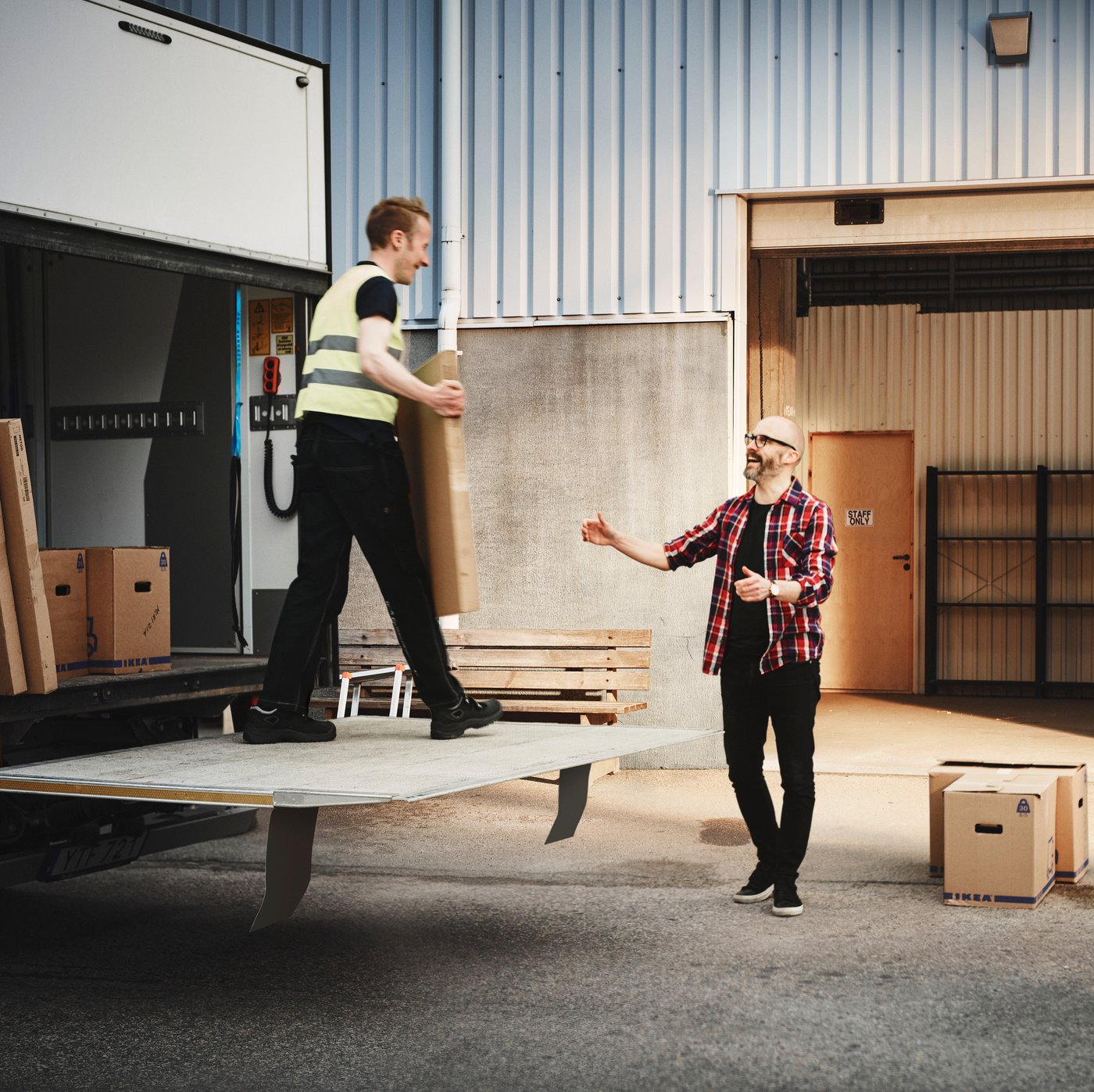 A delivery person standing on the edge of a delivery truck talking to a customer with boxes on the ground.