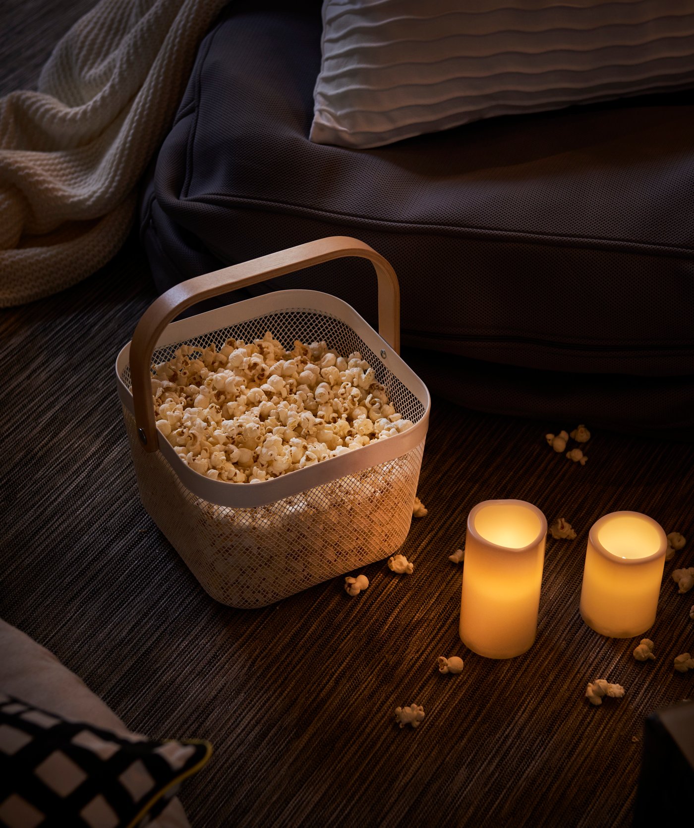 Two battery-operated grey GODAFTON LED block candles next to a RISATORP white basket holding popcorn. 