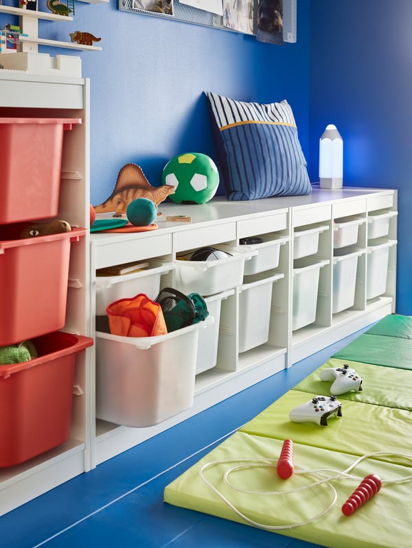 A blue wall with white toy storage organisers, a blue and orange pillow, a dinosaur toy and a green play mat on the floor.