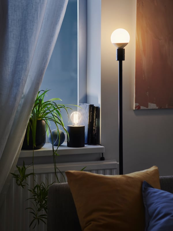 Two lit SOLHETTA LED bulbs, one in a table lamp on a window sill and one in a floorlamp behind a sofa with pillow.