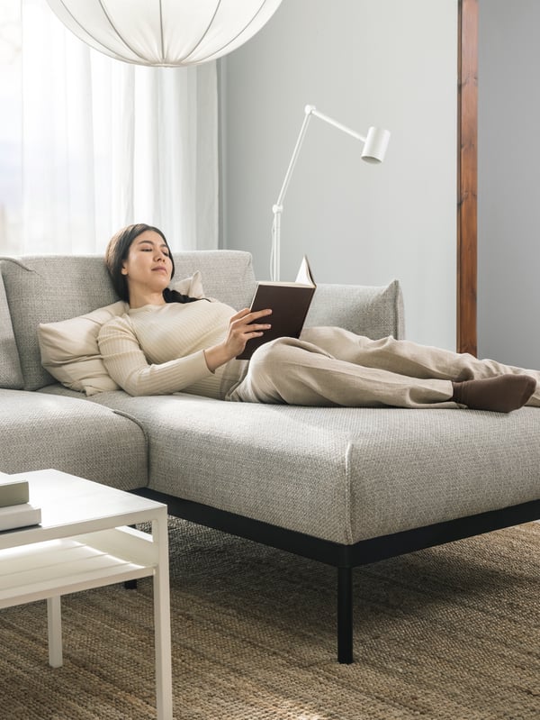 A woman in loungewear reading a book while reclining on a light grey ÄPPLARYD 3-seat sofa with chaise longue.