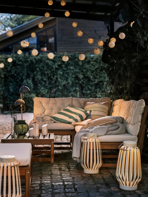 An outdoor lounge area with an outdoor sofa lit by LED lighting chains, four SOMMARLÅNKE floor lamps and LED block candles.