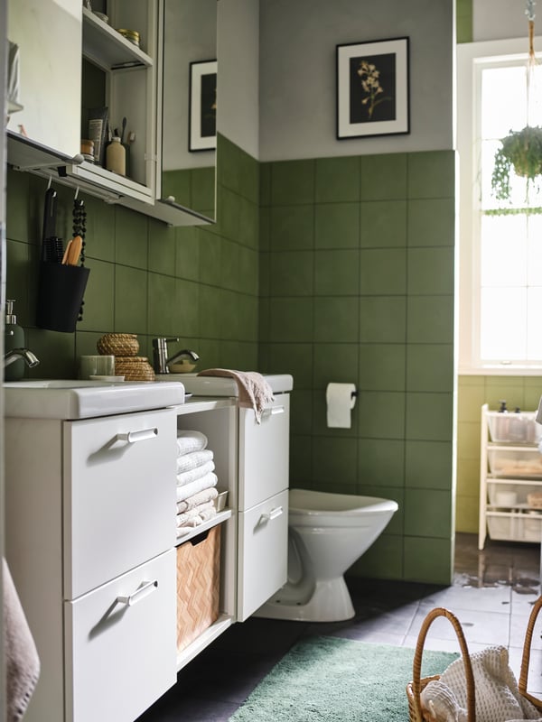 A bathroom with green tiled walls and a mirrored cabinet above two white sinks with drawers and shelves holding white towels.